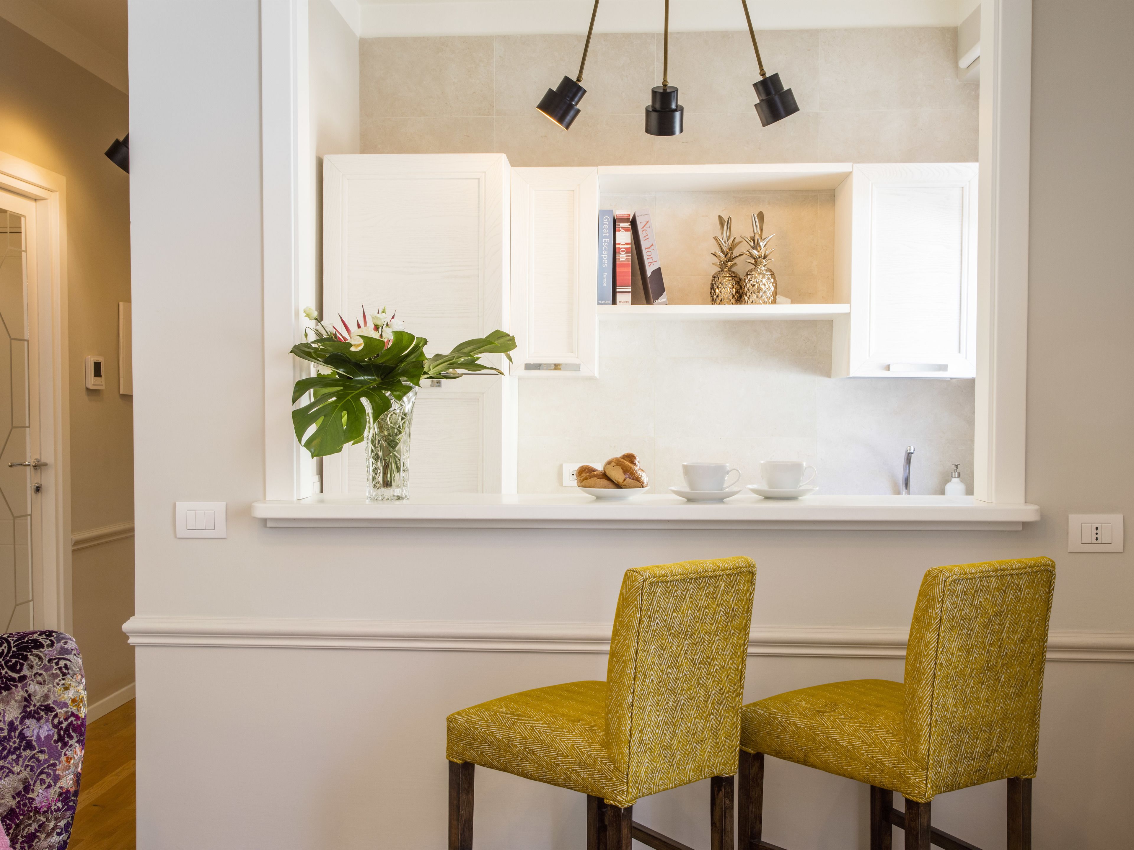 Uma área de refeições com duas cadeiras amarelas, uma mesa branca e uma estante com livros e objetos decorativos.