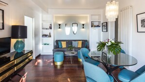 Ein modernes Wohnzimmer mit einer Couch, einem Couchtisch und Sesseln. Ein Fernseher, eine Lampe und ein Kronleuchter sind ebenfalls vorhanden.
