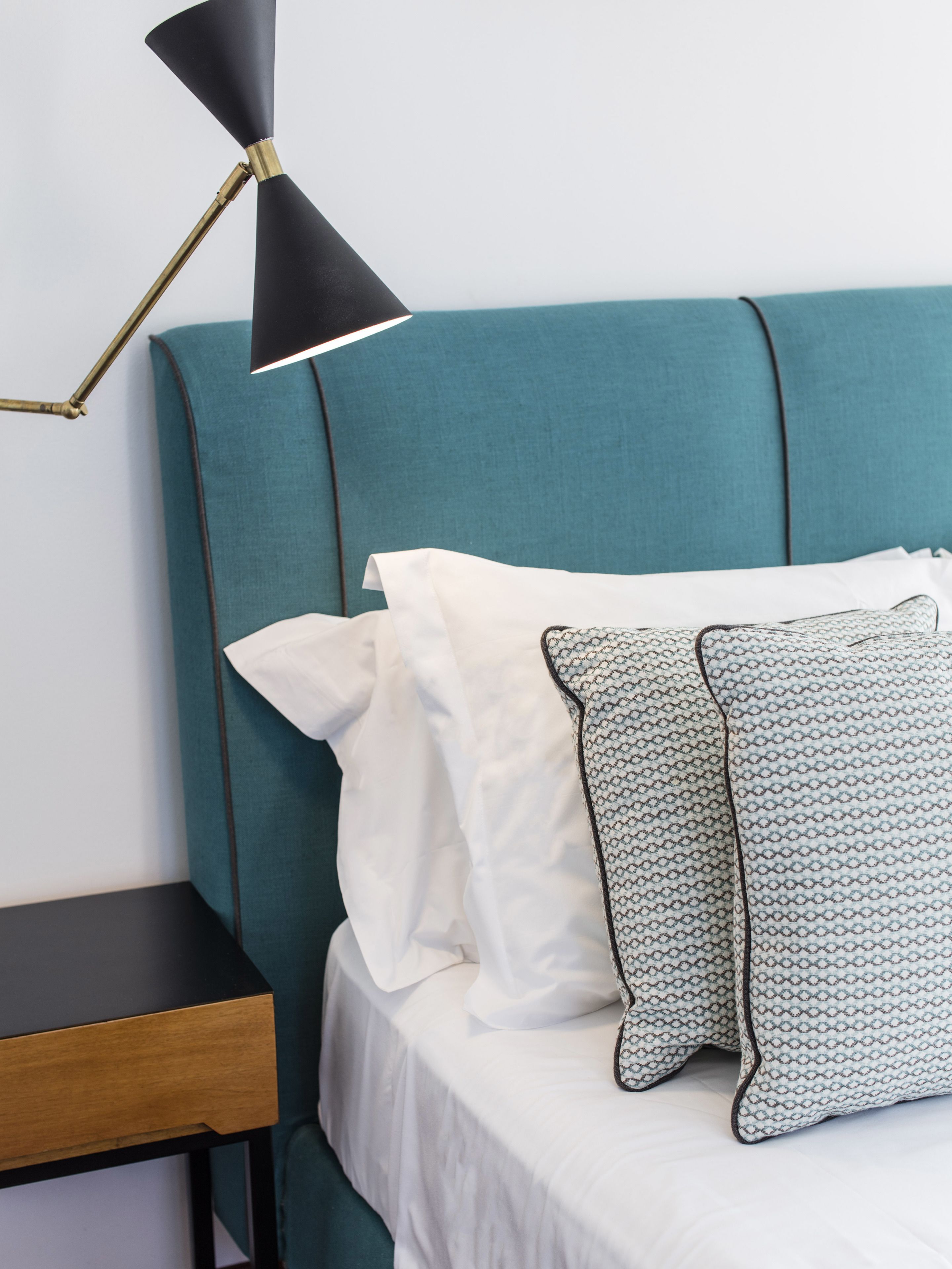 Un lit avec une tête de lit turquoise, une literie blanche et des coussins à motifs. Une lampe de bureau noire avec un pied doré est posée sur le côté.