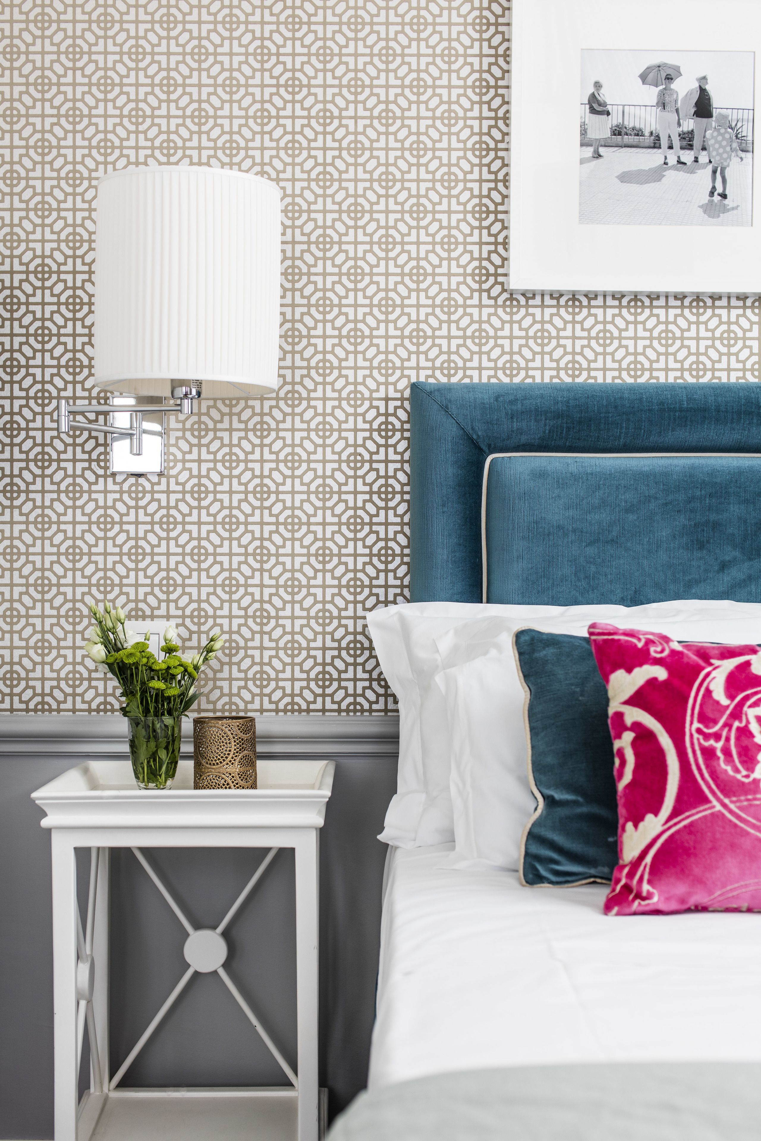 A neatly arranged bedroom with a patterned wallpaper, a white bedside table with a vase of flowers, a blue headboard, and a framed picture on the wall.