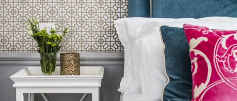 A neatly arranged bedroom with a patterned wallpaper, a white bedside table with a vase of flowers, a blue headboard, and a framed picture on the wall.