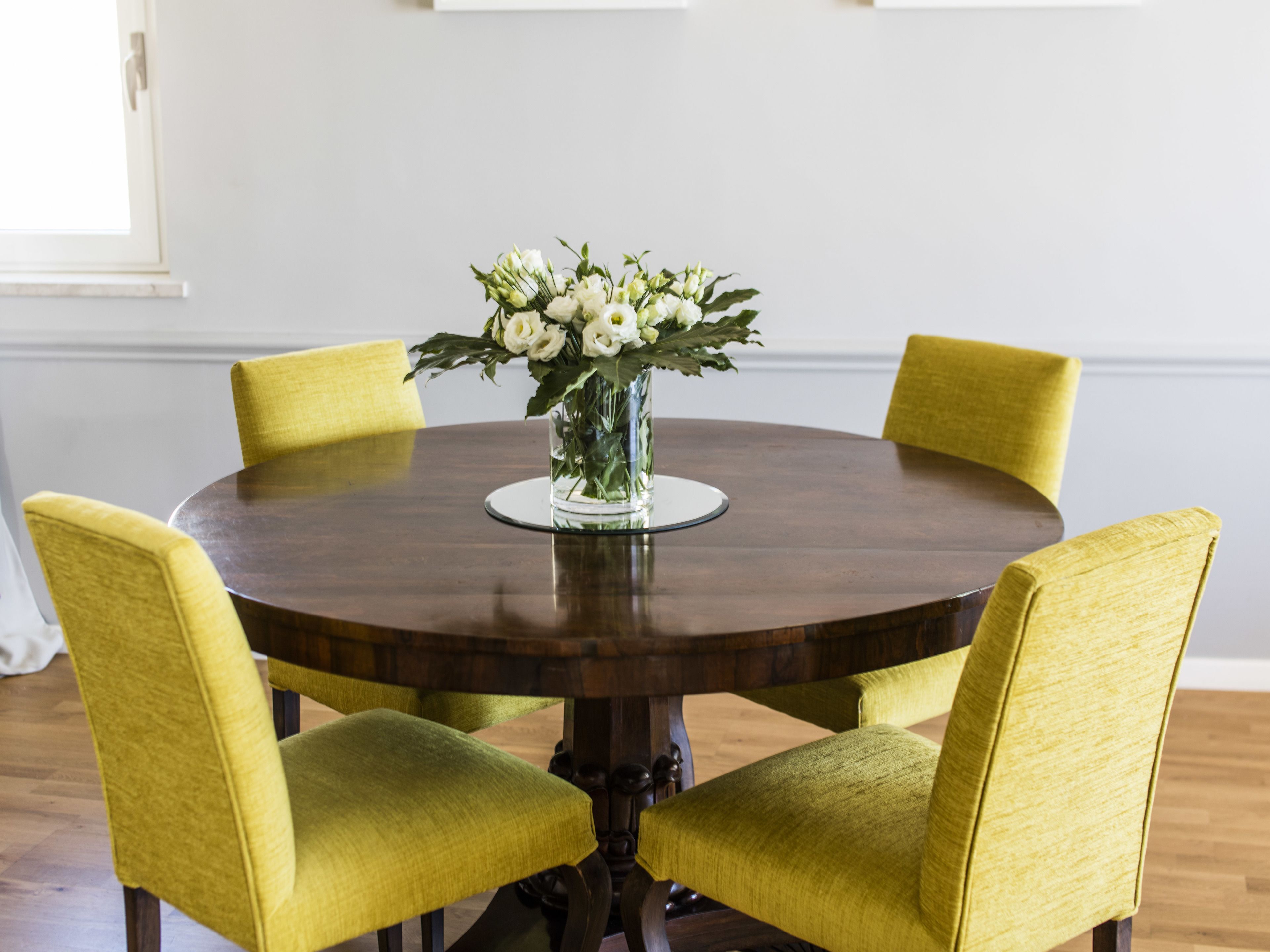 A round wooden dining table with a glass vase of white flowers in the center, surrounded by four yellow chairs.