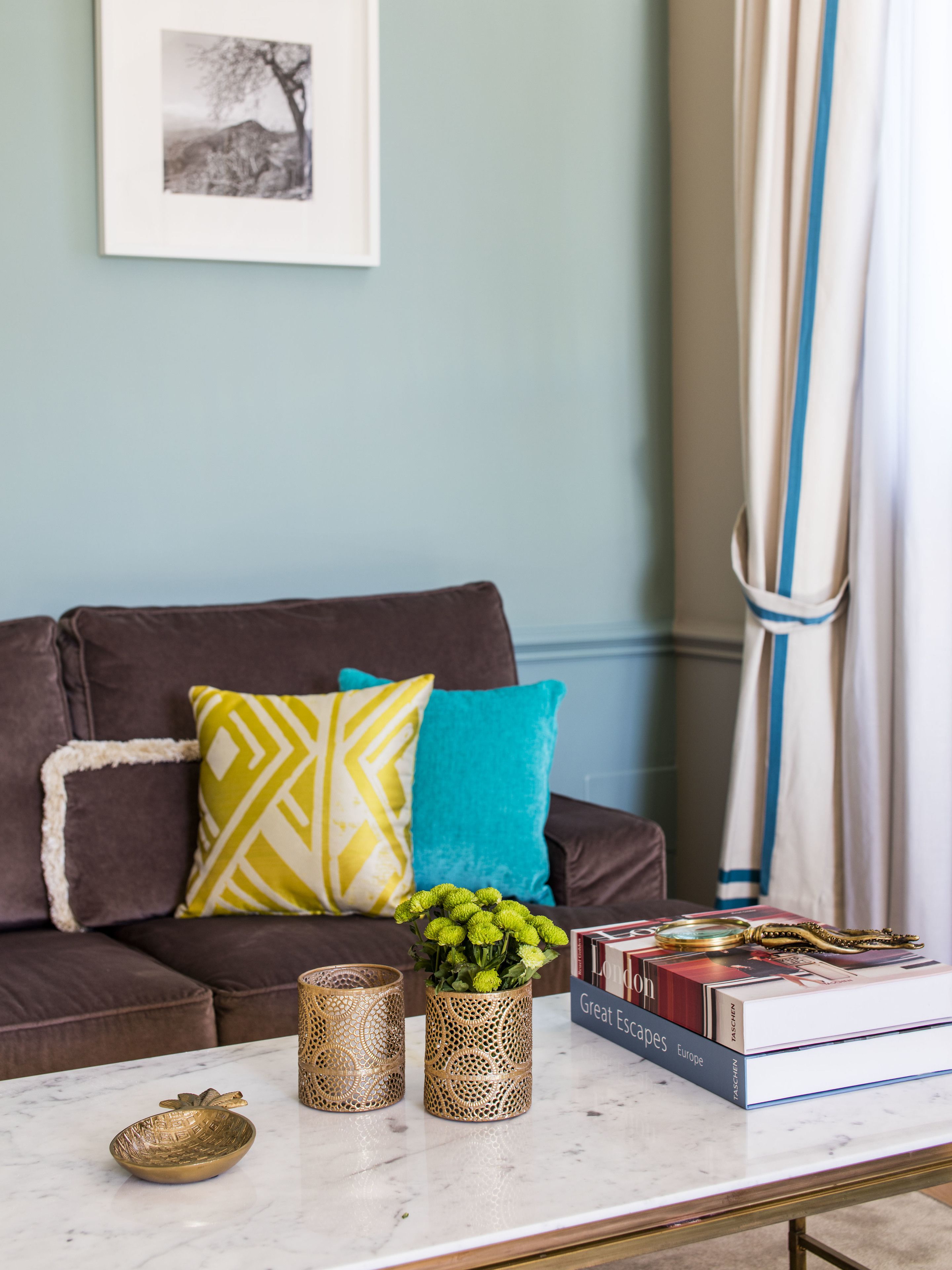 Una sala de estar con un sofá marrón, un cojín verde y amarillo estampado, un jarrón con flores verdes y una pila de libros sobre una mesa de centro de mármol.