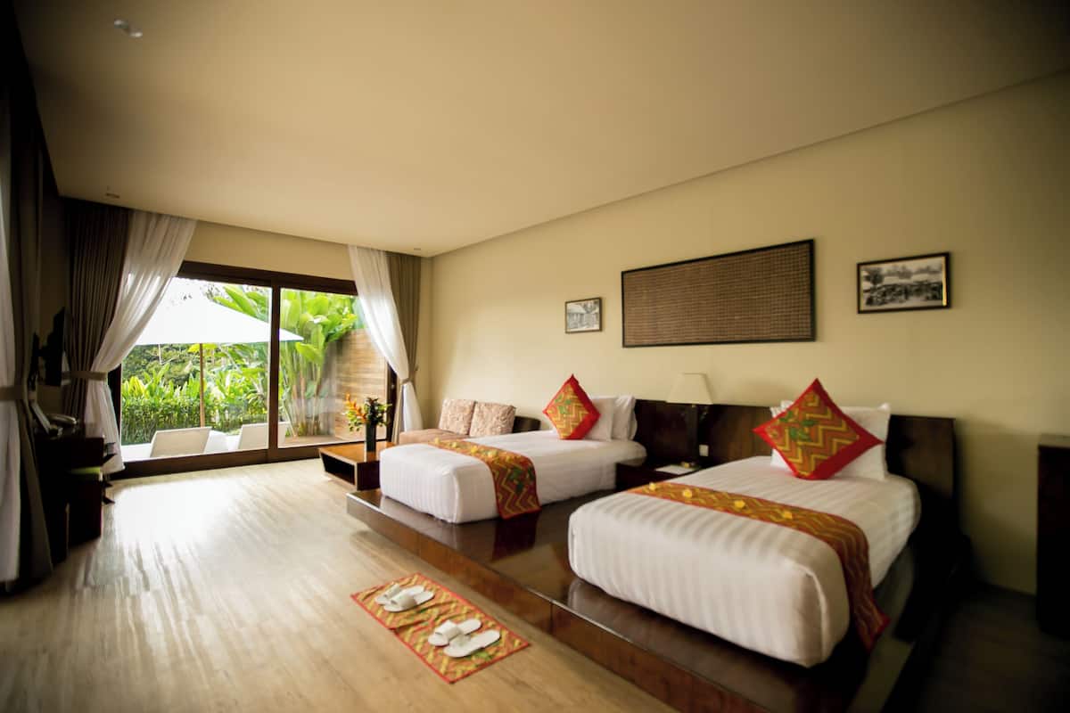 A hotel room with two beds, a large window with curtains, a wooden floor, and a view of greenery outside.