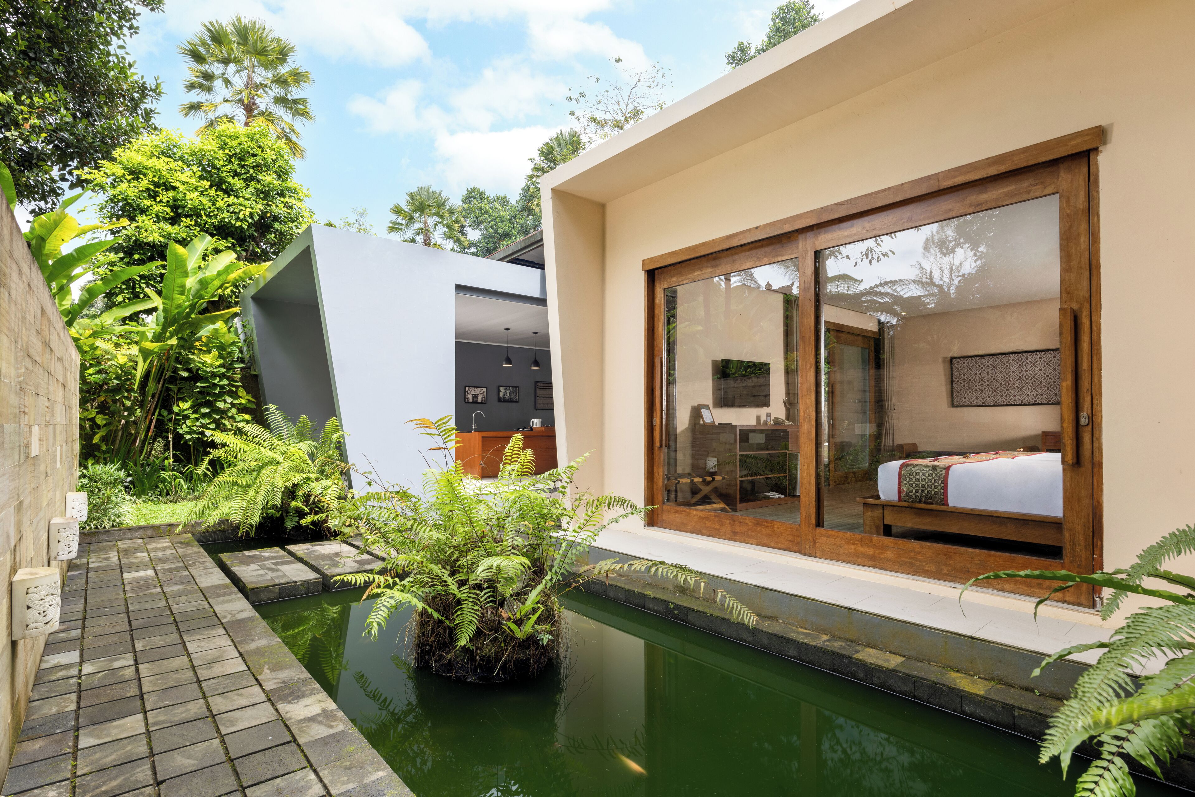 A modern villa with a swimming pool, a large sliding glass door leading to a bedroom, and lush greenery surrounding the property.