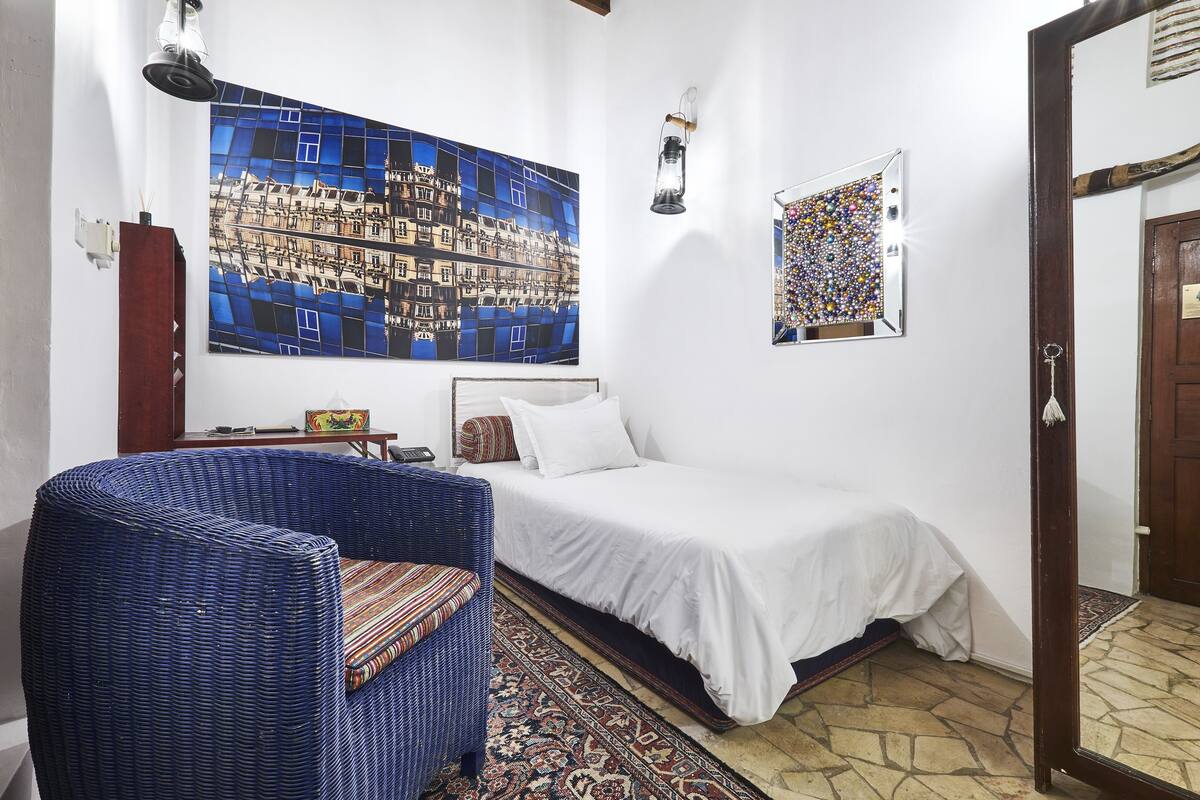 Ein Zimmer mit einem Bett, einem Korbstuhl, einem großen Spiegel und einer Wand mit zwei Kunstwerken.