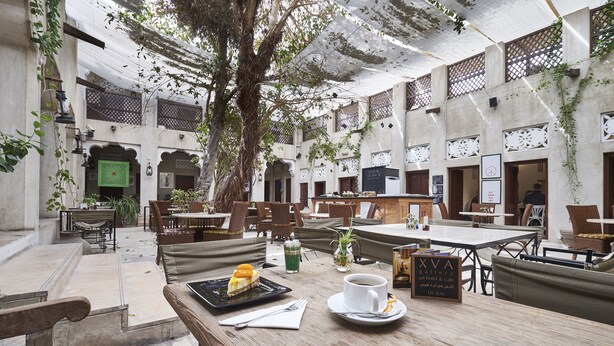 Um pátio com mesas e bancos de madeira, uma árvore e uma placa com a inscrição 'XVA Gallery art hotel & cafe Dubai'.