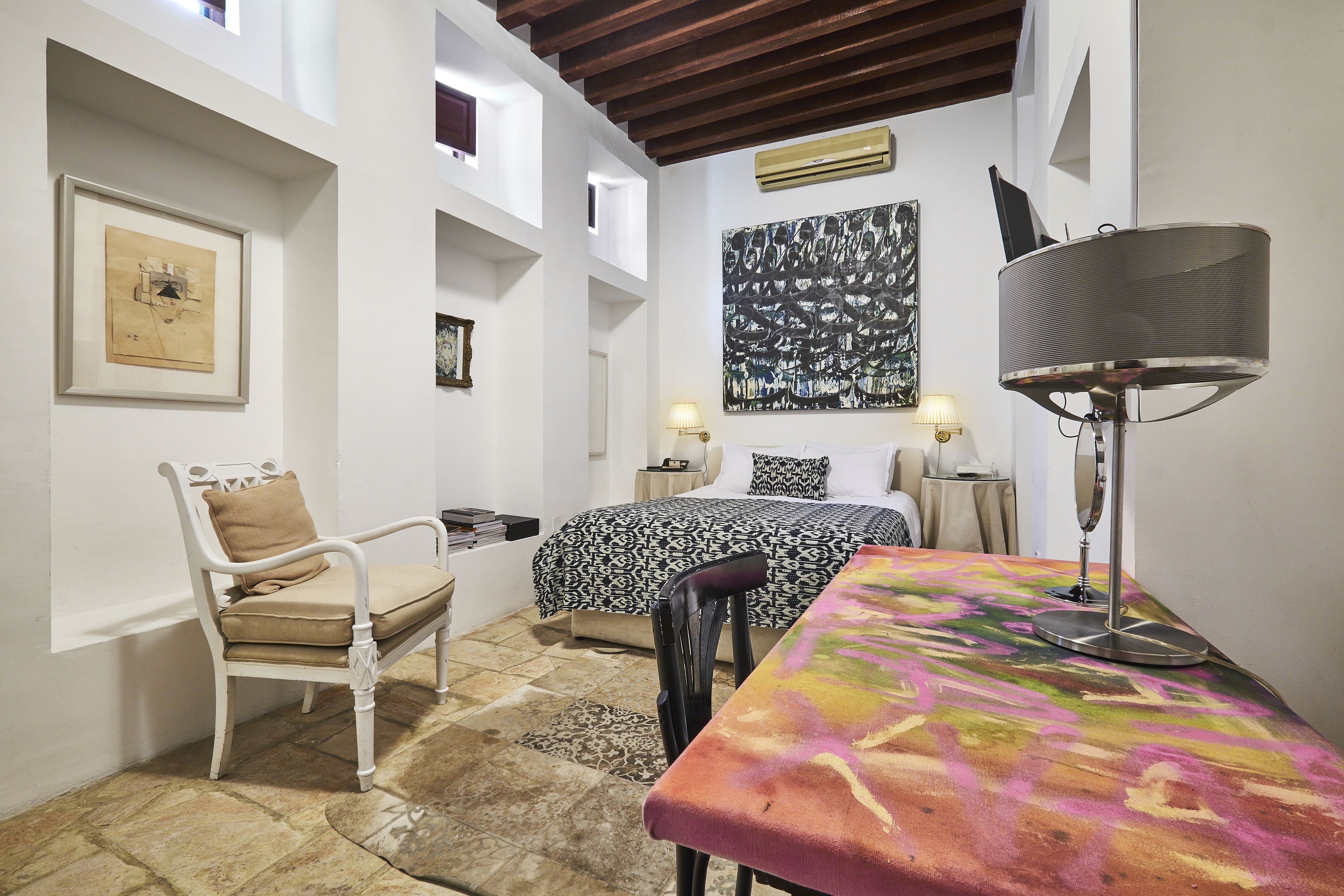Una stanza arredata con un letto, una sedia, un tavolo con una tovaglia colorata, una lampada e opere d'arte alle pareti.