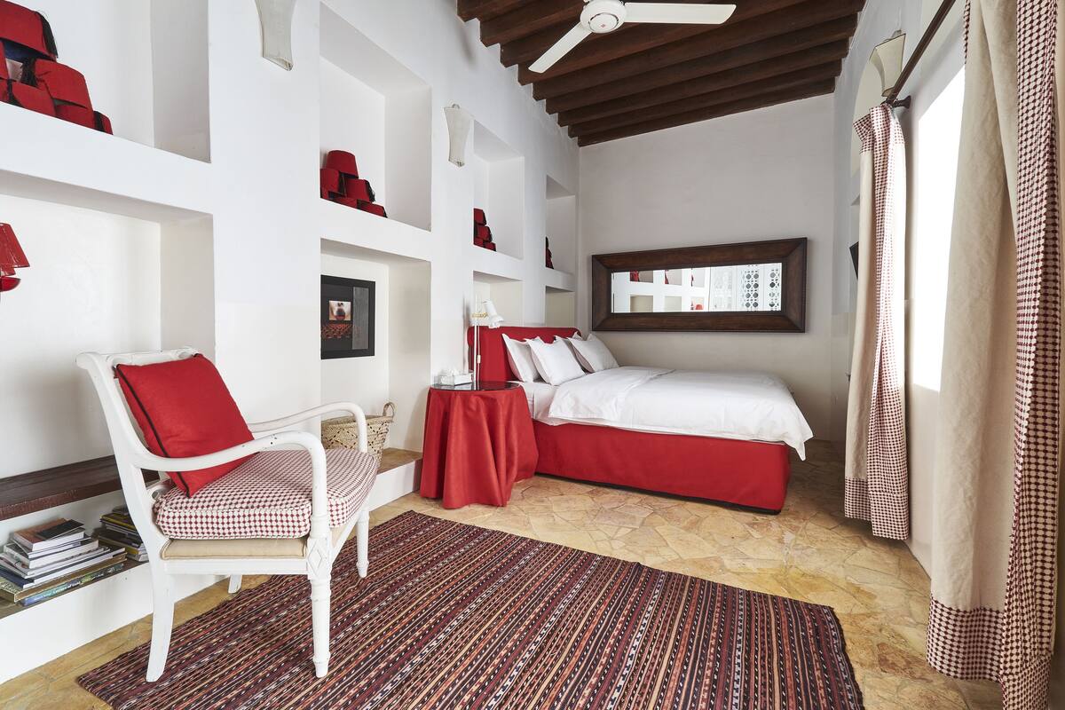 Ein Schlafzimmer mit einem Bett, einem Sessel, einem kleinen Tisch und einer Ablage mit roten Gegenständen.