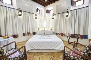 A bedroom with a large bed, two bedside lamps, a small table with fruit, and patterned chairs.