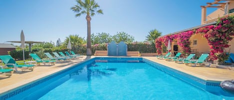A swimming pool with clear blue water, surrounded by lounge chairs and a pergola with flowering plants.