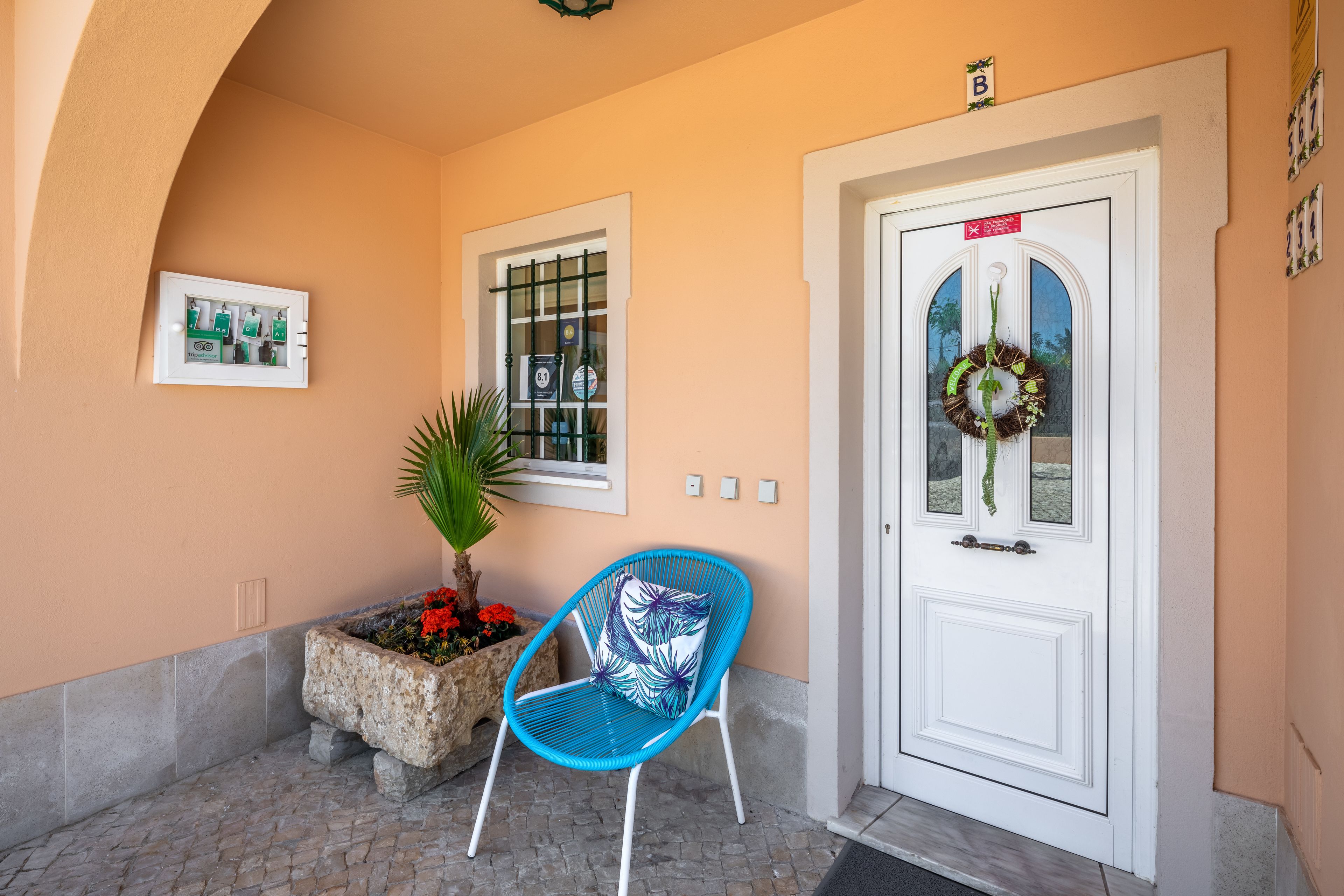 A white door with a wreath, a blue chair, a stone planter with red flowers, and a framed picture on the wall.