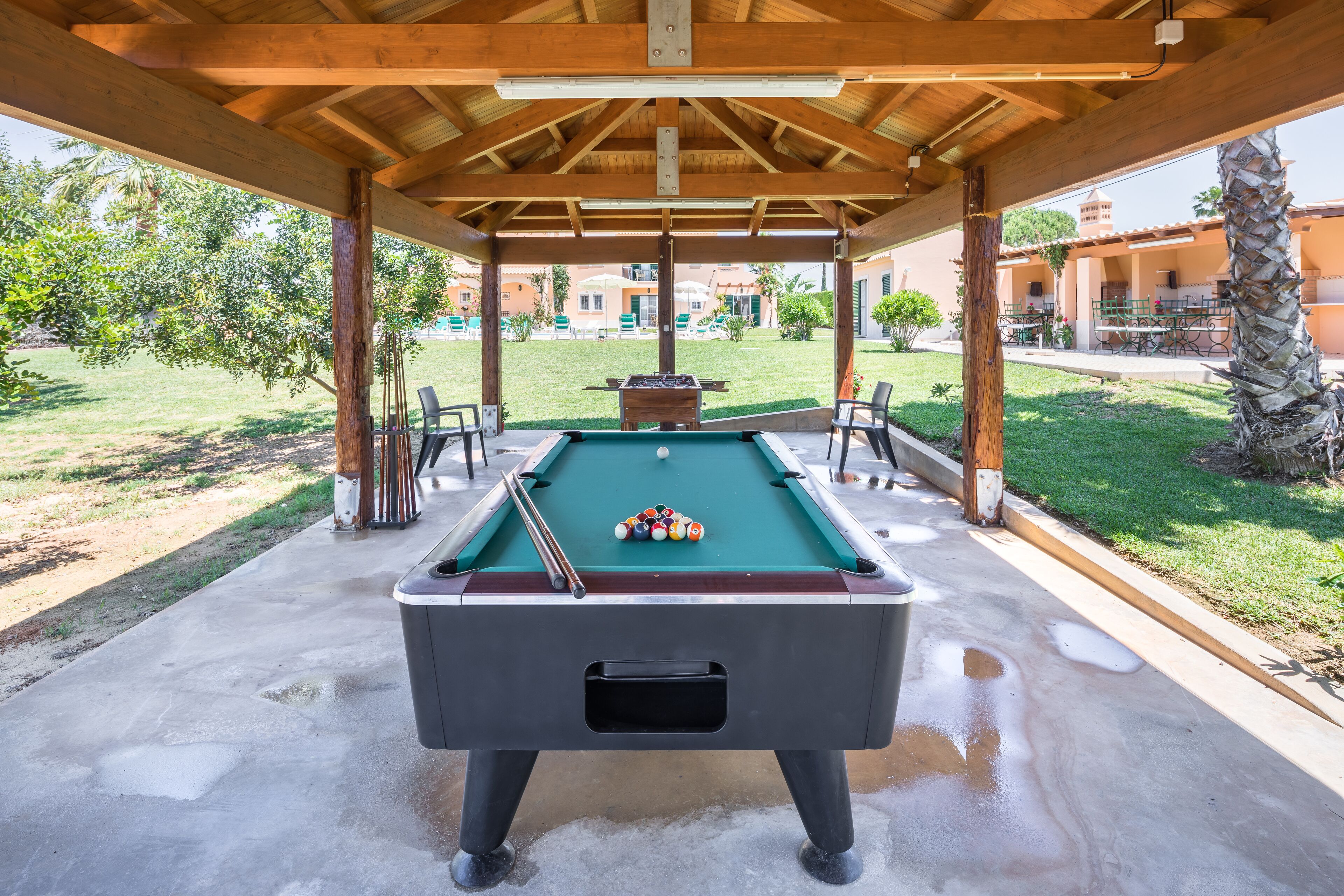 A pool table under a wooden pavilion with a view of a pool area and buildings in the background.