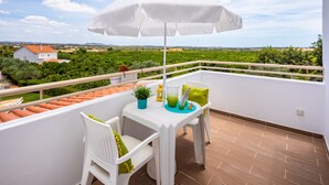 A balcony with a white table, chairs, and a white umbrella. There is a view of greenery and houses in the background. - Flôr da Laranja, Albufeira (Albufeira)