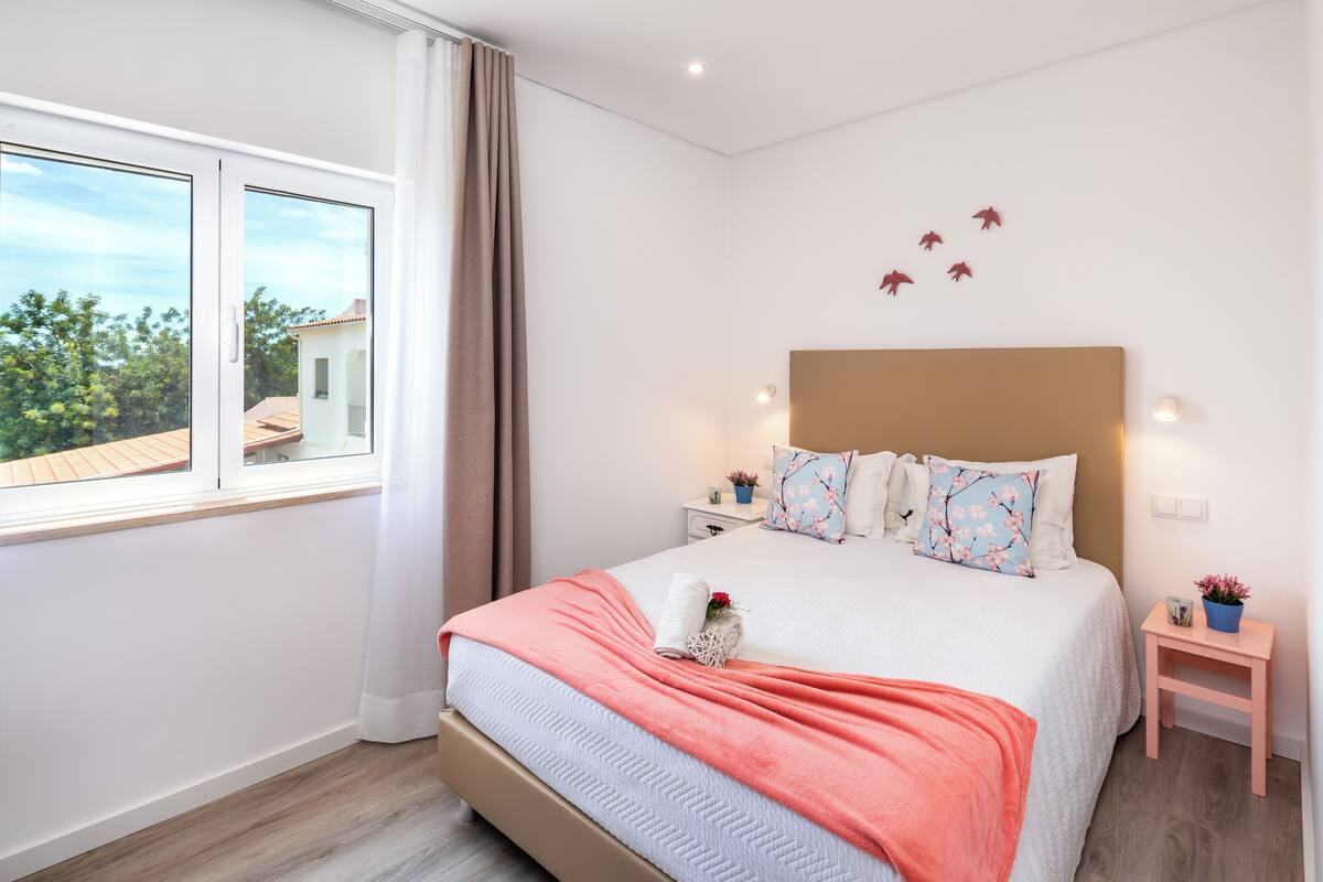 A bedroom with a large bed, a nightstand, a small table with a plant, and a window with curtains.