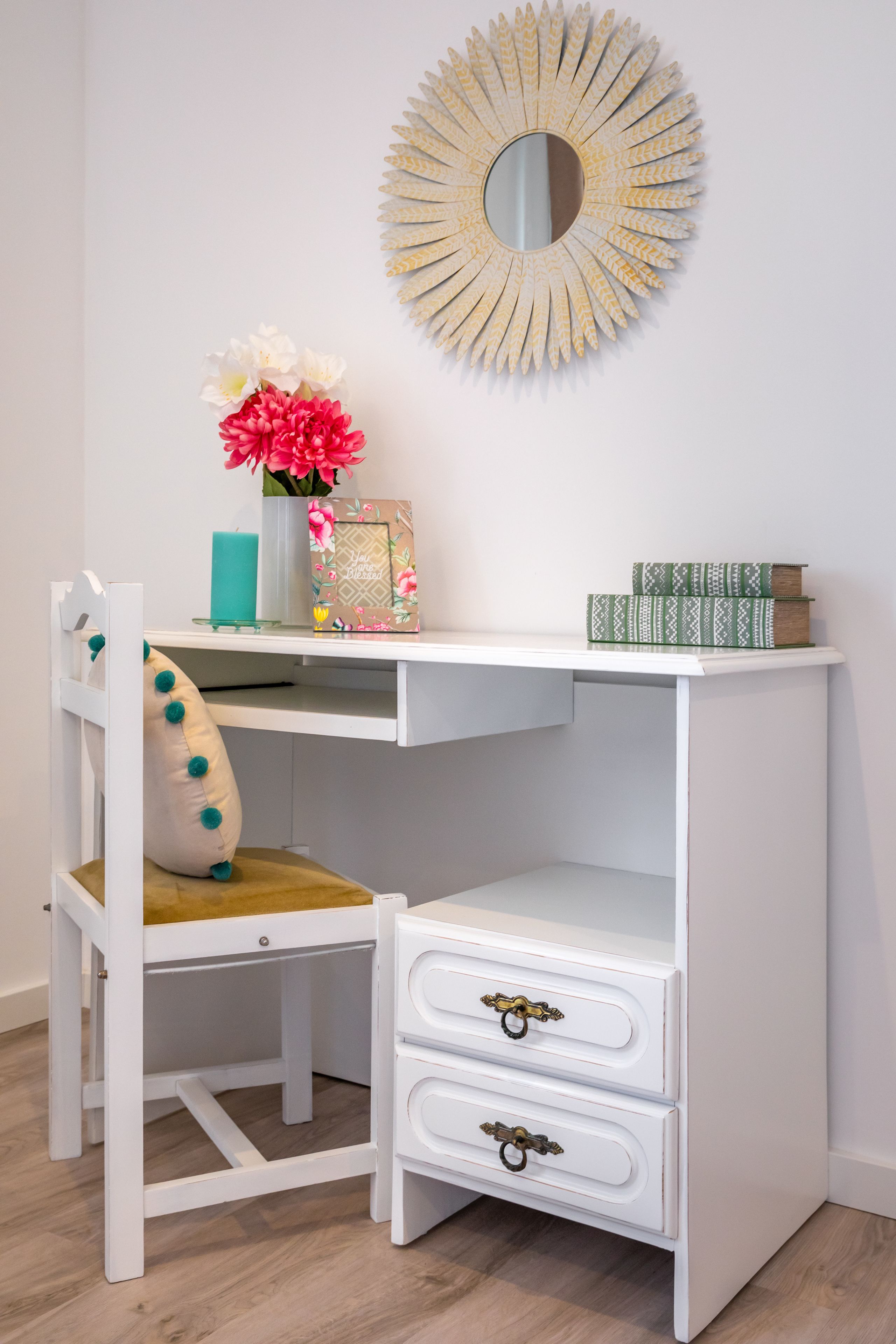 A white desk with drawers, a chair with a wooden seat and teal accents, a mirror with a sunburst frame, and a vase with flowers.