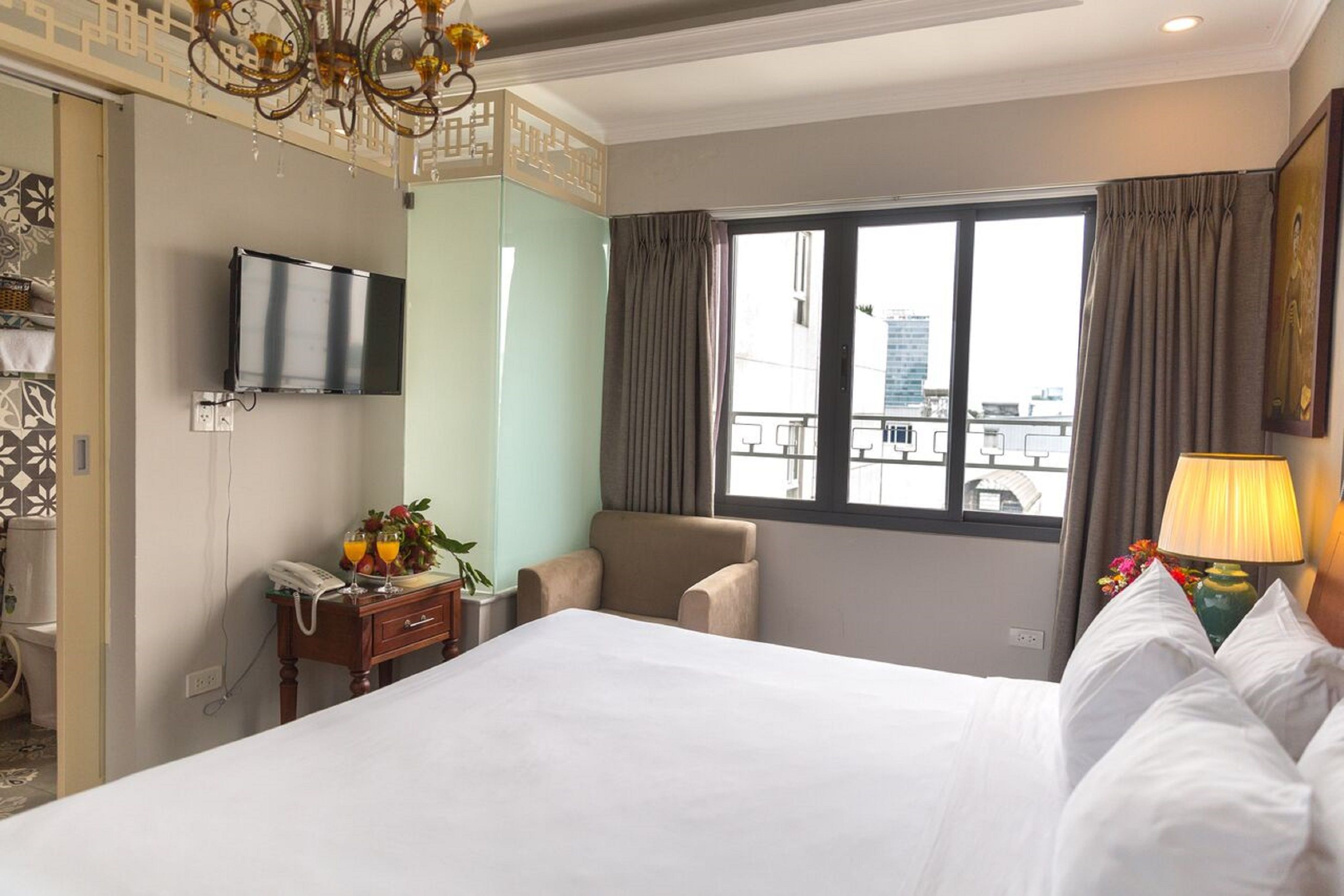 Ein Hotelzimmer mit einem Bett, einem an der Wand montierten Fernseher, einem Sessel, einem Schreibtisch mit Telefon und einer Blumenvase sowie einem Fenster mit Vorhängen.