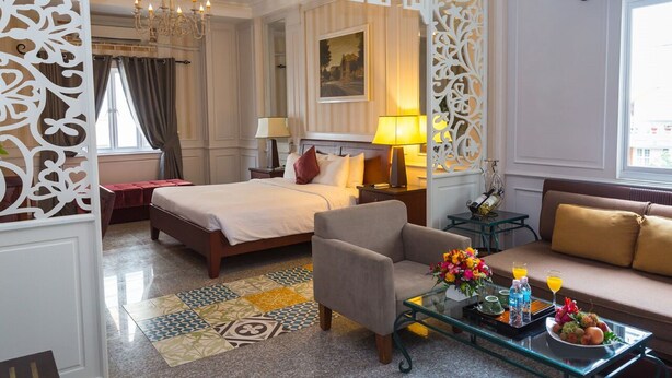 Ein Hotelzimmer mit Bett, Sofa, einem Glastisch mit Obst und Getränken, einem dekorativen Raumteiler und einer Chandelier.