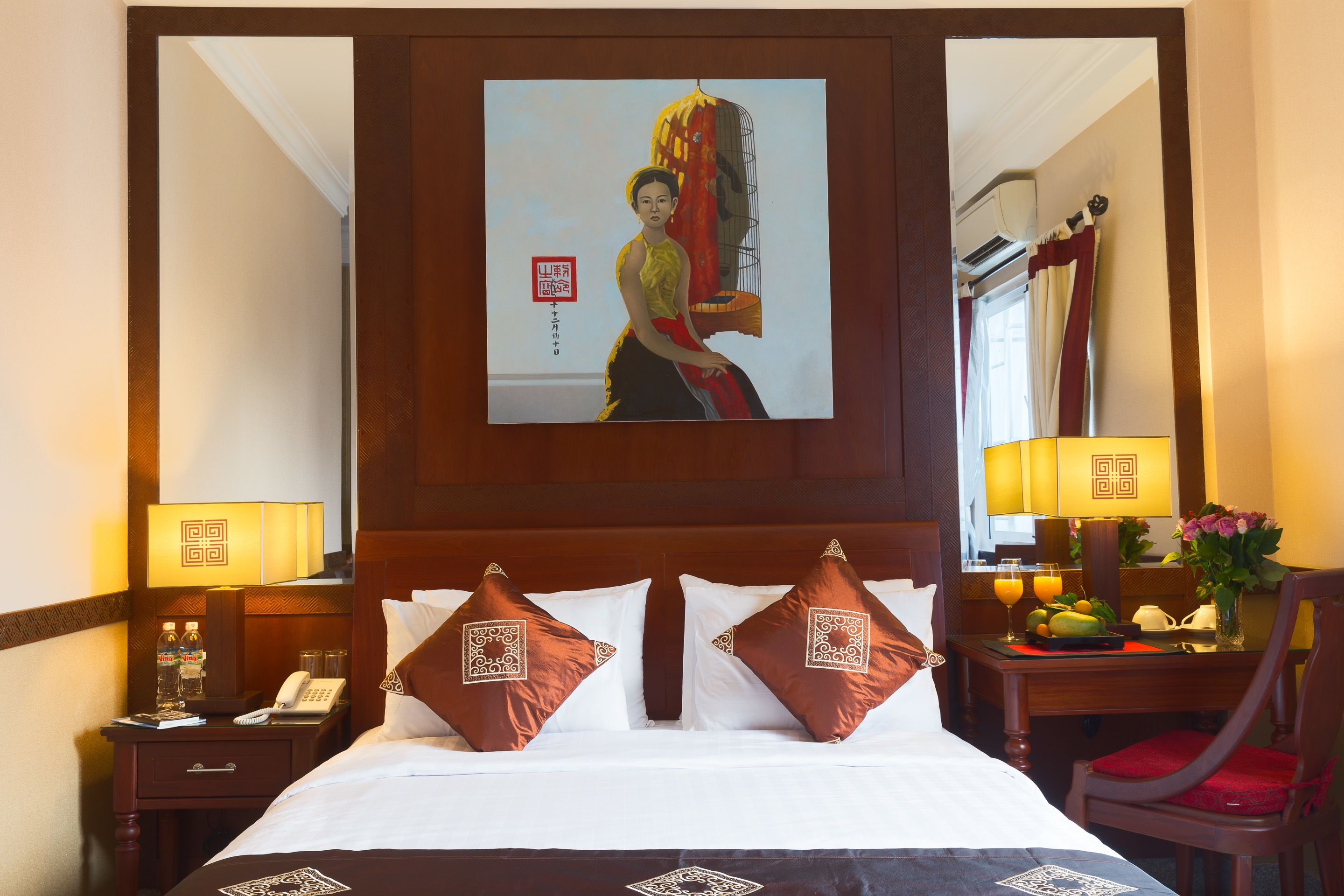 A hotel room with a bed, two pillows with a geometric design, a nightstand with a phone and a lamp, a wooden headboard, and a painting above the bed.