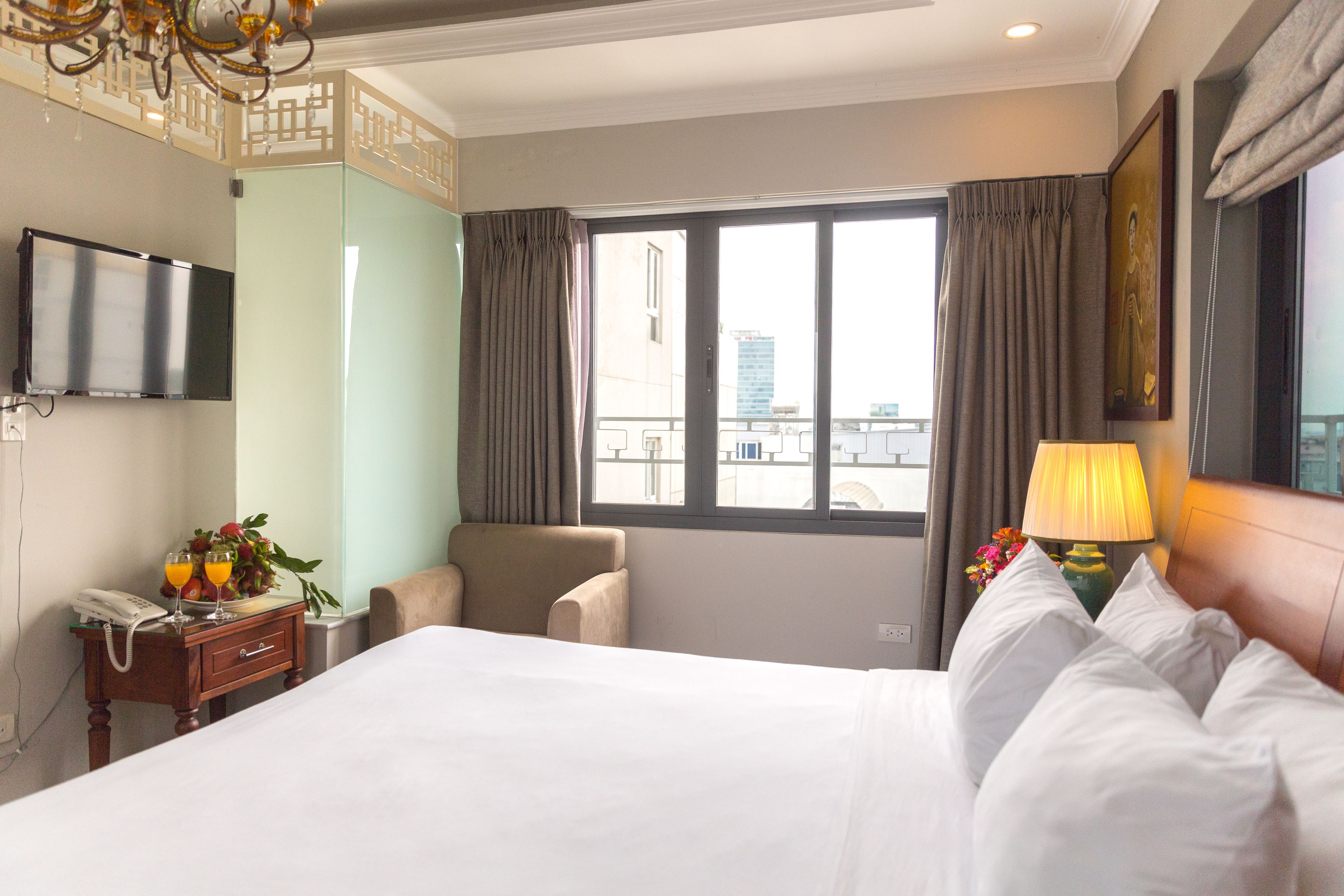 Una habitación de hotel con cama, sillón, televisor, ventana con cortinas y lámpara.