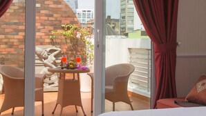 Ein Hotelzimmer mit einem Bett, einem Balkon mit Korbstühlen, einem Tisch mit Getränken und Blumen und Blick auf hohe Gebäude.