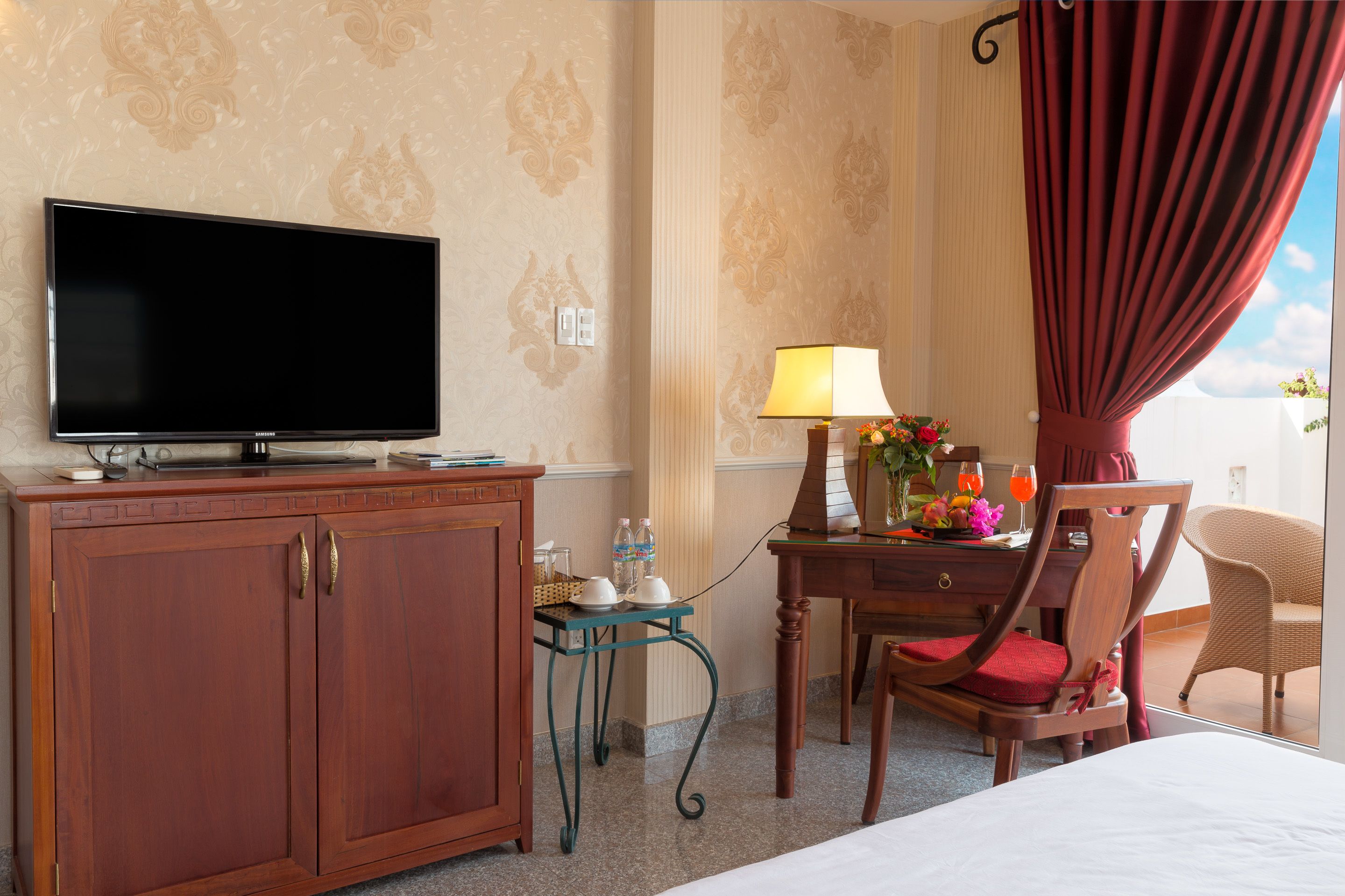 A hotel room with a flat-screen TV, a wooden cabinet, a small table with a lamp and flowers, a chair, and a bed.