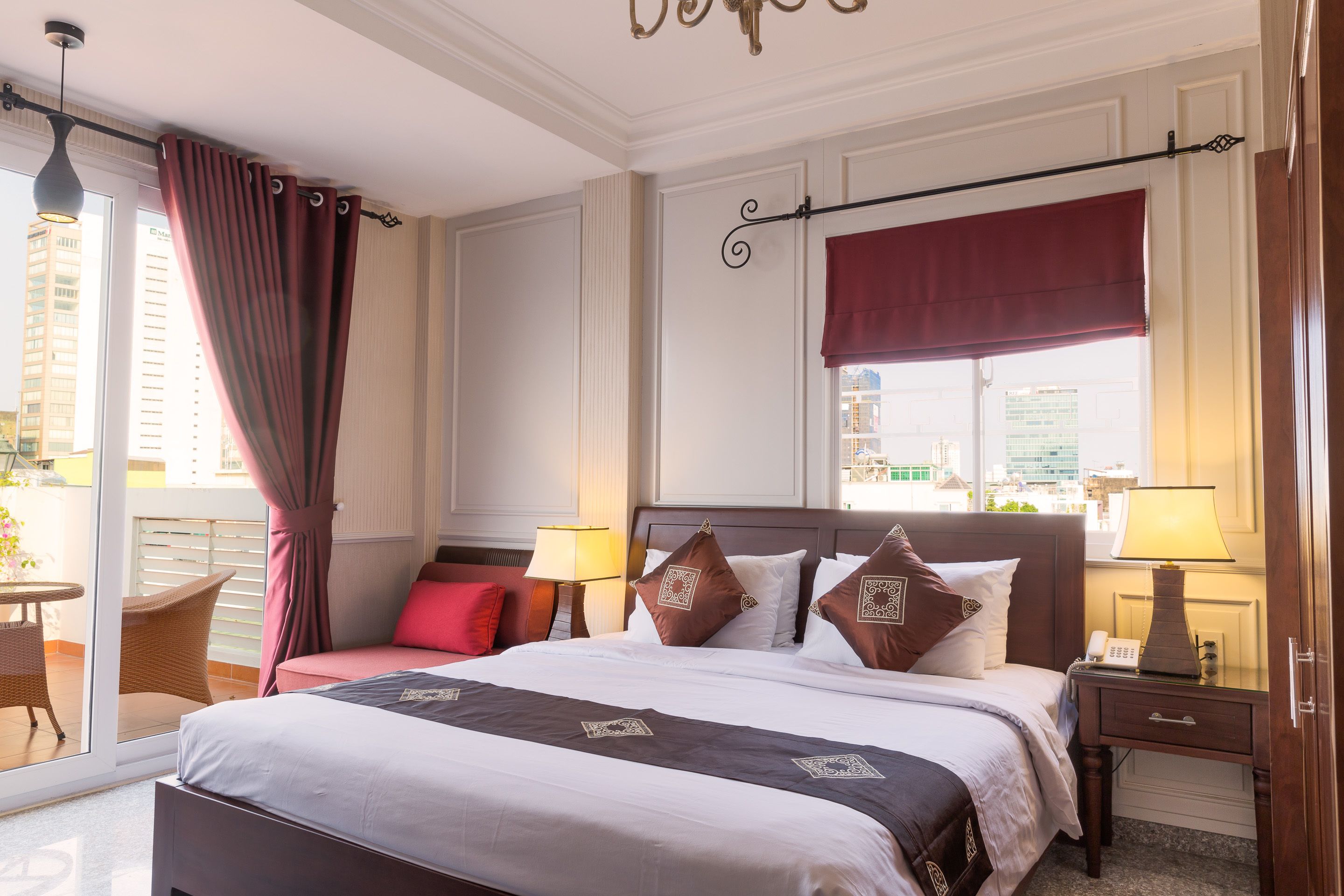 Een hotelkamer met een groot bed, twee nachtkastjes met lampen, een raam met een rode vouwgordijn en een balkon met rieten meubels.