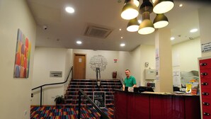 Un hall de hotel con un mostrador de recepción, una escalera con alfombra de colores y una pared con un cartel de nube de palabras.