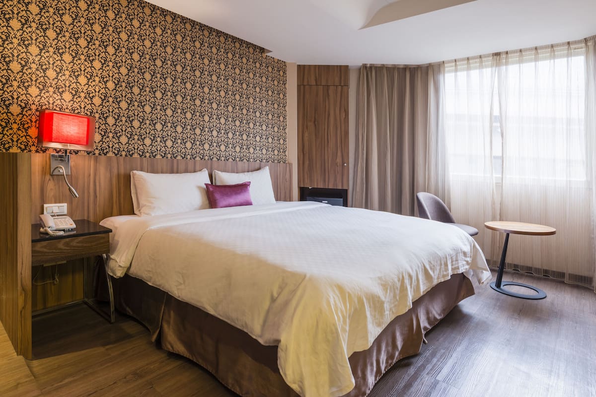 Hotellihuone, jossa on suuri sänky, yöpöytä lampun kanssa, tuoli ja näkymä ulos ohuiden verhojen läpi.
