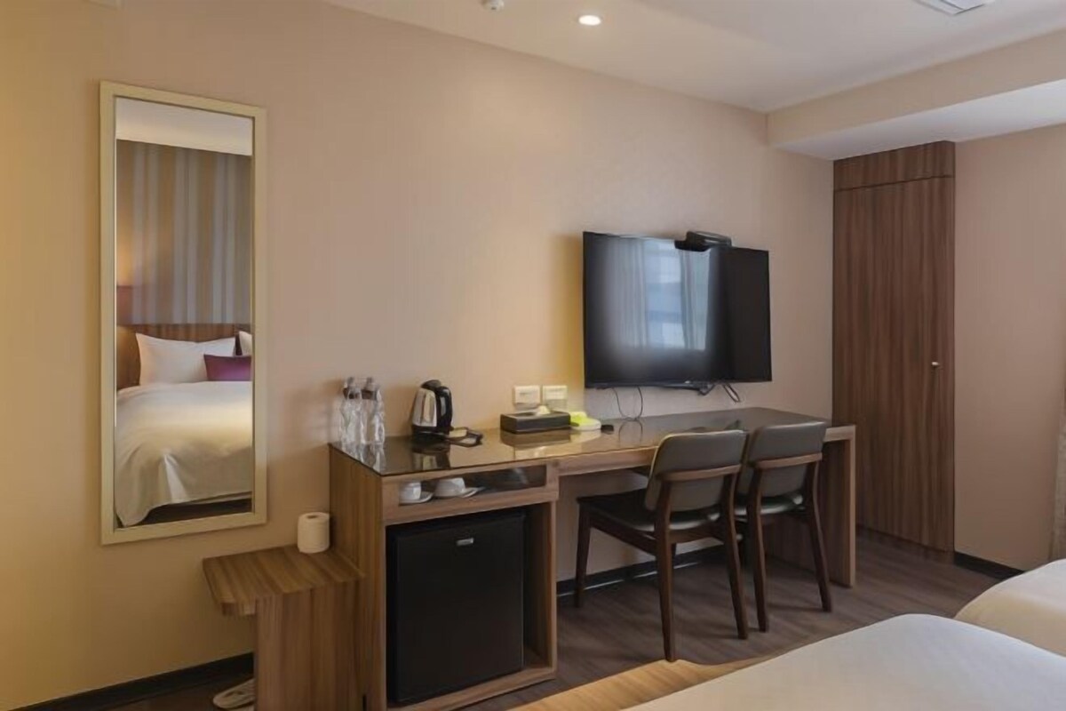 A hotel room with a bed, a desk with a chair, a television, a small refrigerator, and a mirror.