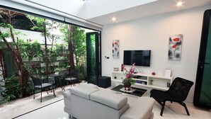 A modern living room with a grey sofa, black chairs, a TV, and a glass door leading to a garden. - Villa Labaron (Koh Samui)