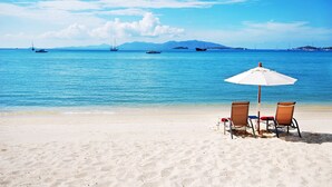 A beach with two chairs and a white umbrella, a clear blue sea, and boats in the distance. - Villa Labaron (Koh Samui)