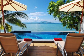 Un espace aménagé au bord de la piscine, avec deux chaises longues et des parasols, d’où l’on peut admirer une mer bleue et limpide où naviguent des bateaux.
