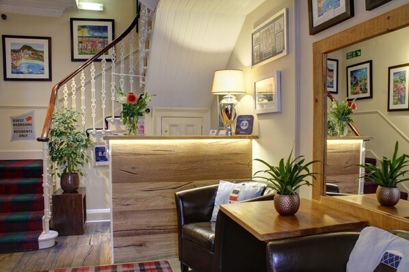 A hotel lobby with a wooden reception desk, a black leather sofa, a staircase with a white railing, and several framed pictures on the walls. - No.12 Hotel & Bistro (North Berwick)