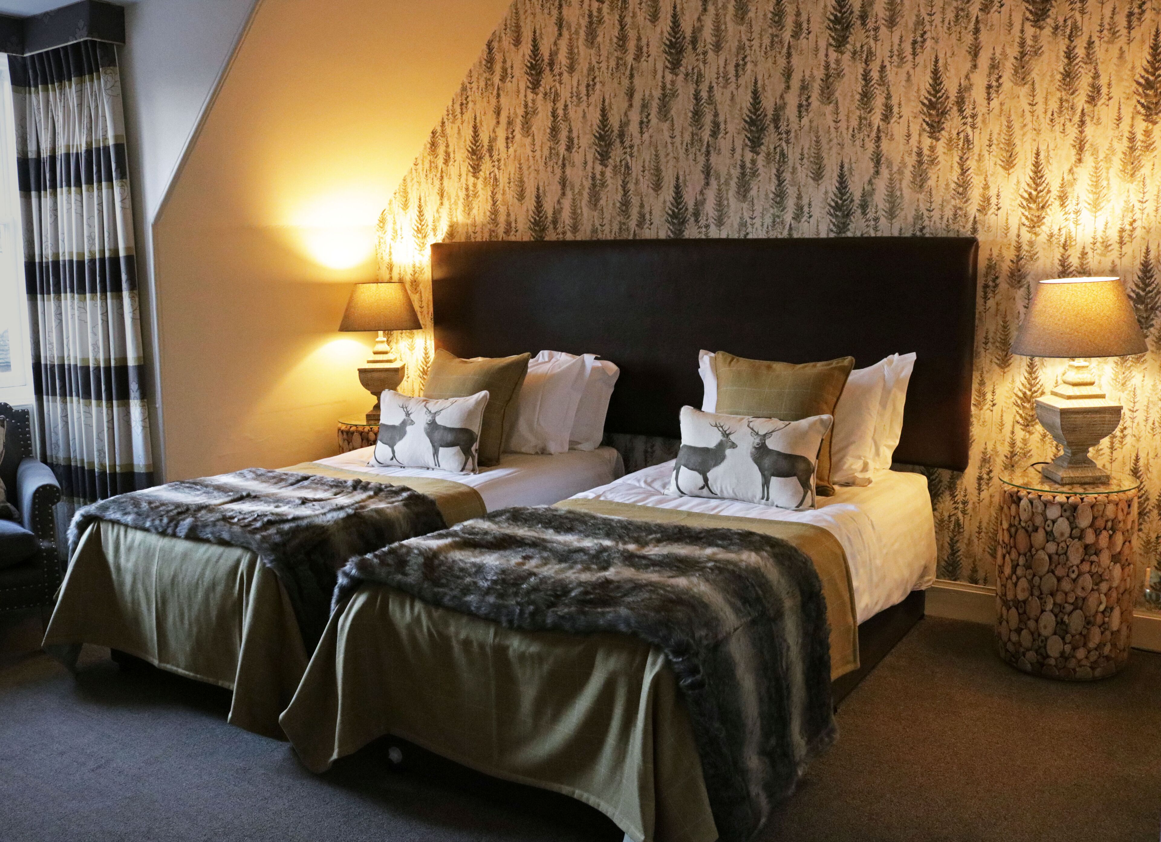 A hotel room with two beds, a large dark headboard, and a forest-themed wallpaper. There are two bedside lamps and a small table with a lamp.