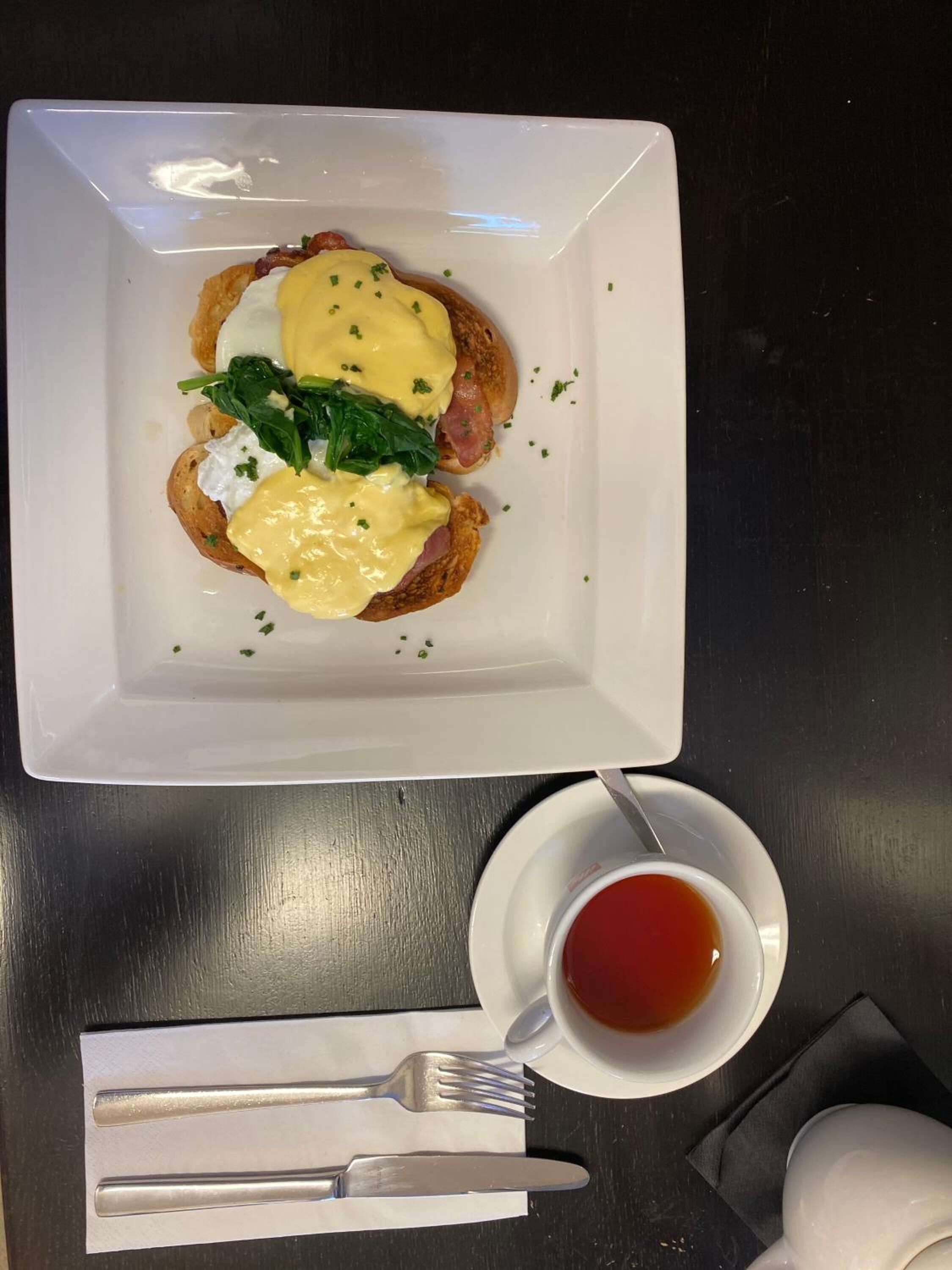 A breakfast plate with eggs benedict, a cup of tea, and cutlery on a table.