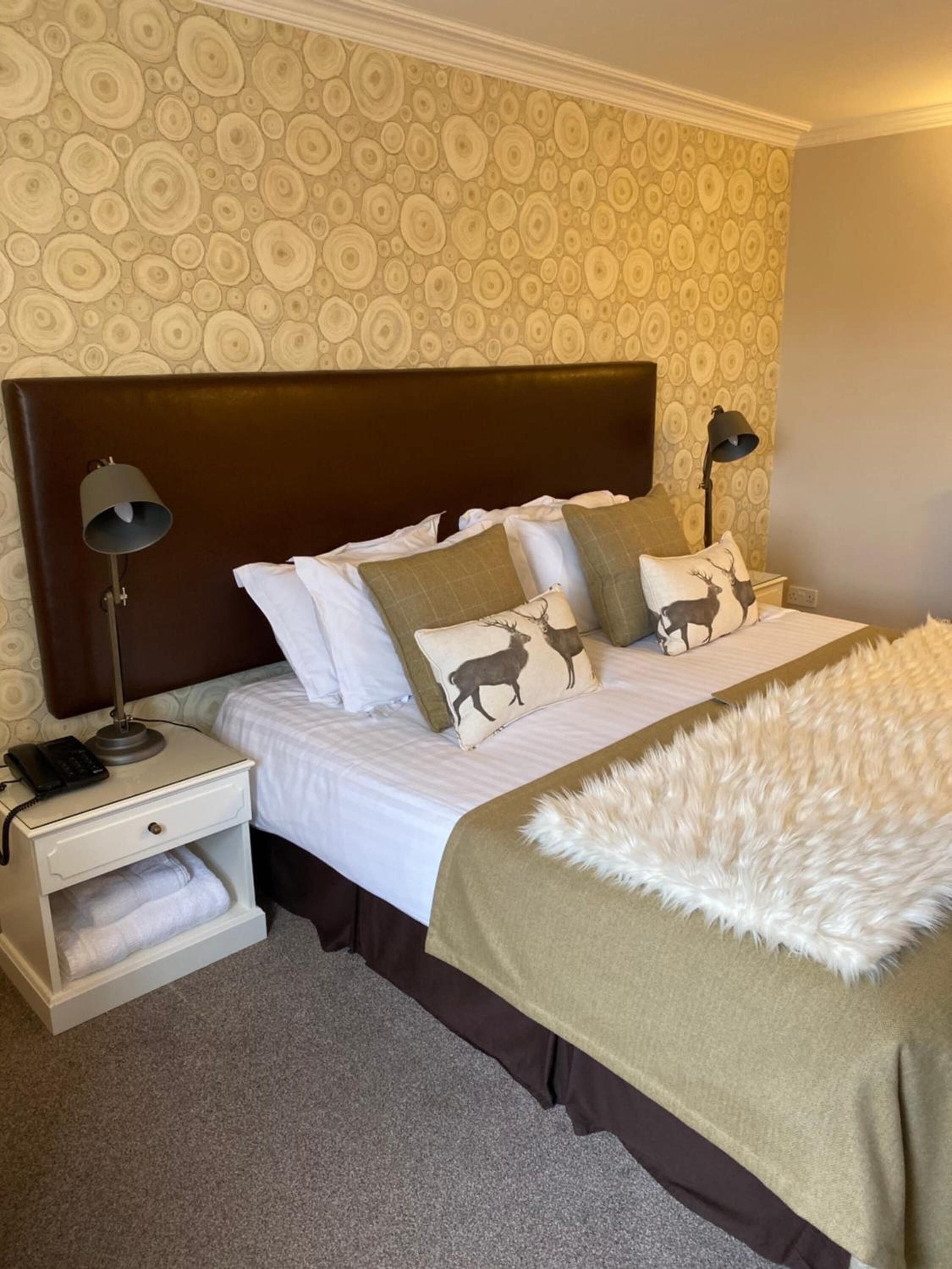 Un lit bien fait, avec une tête de lit marron, du linge de lit blanc et des coussins décoratifs, dont un orné d’un motif de cerf. On y trouve une table de chevet blanche avec une lampe et un téléphone, ainsi qu’une couverture blanche et moelleuse posée sur le lit.