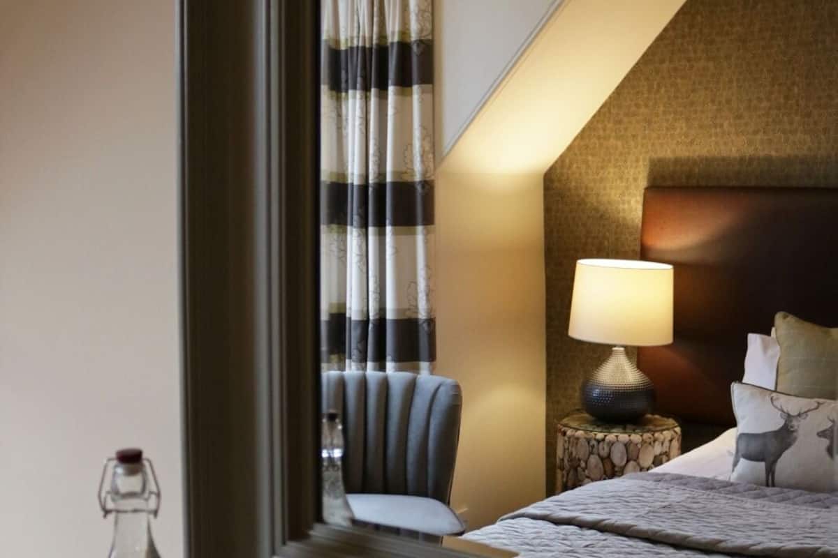 A hotel room with a large mirror reflecting a bed, a nightstand with a lamp, a chair, and a decorative pillow.