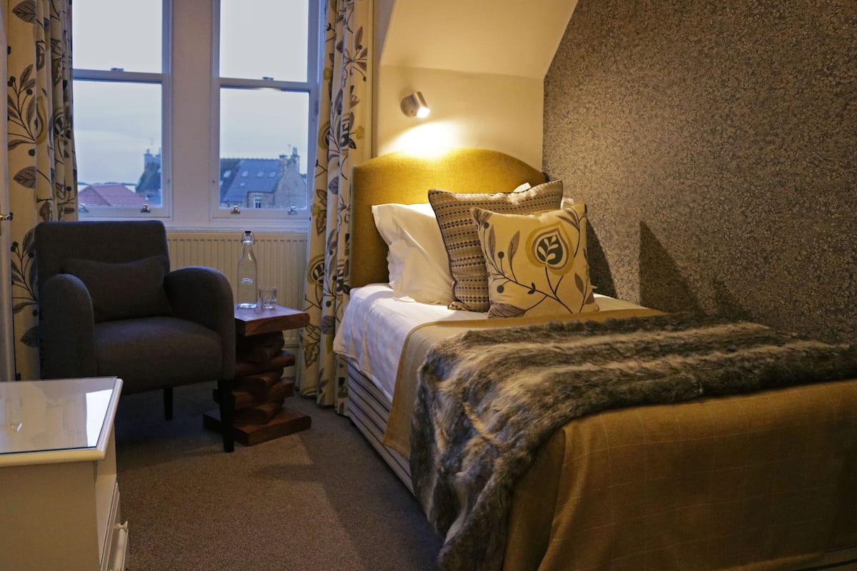 A bedroom with a bed, a chair, a bedside table, and a window with curtains.