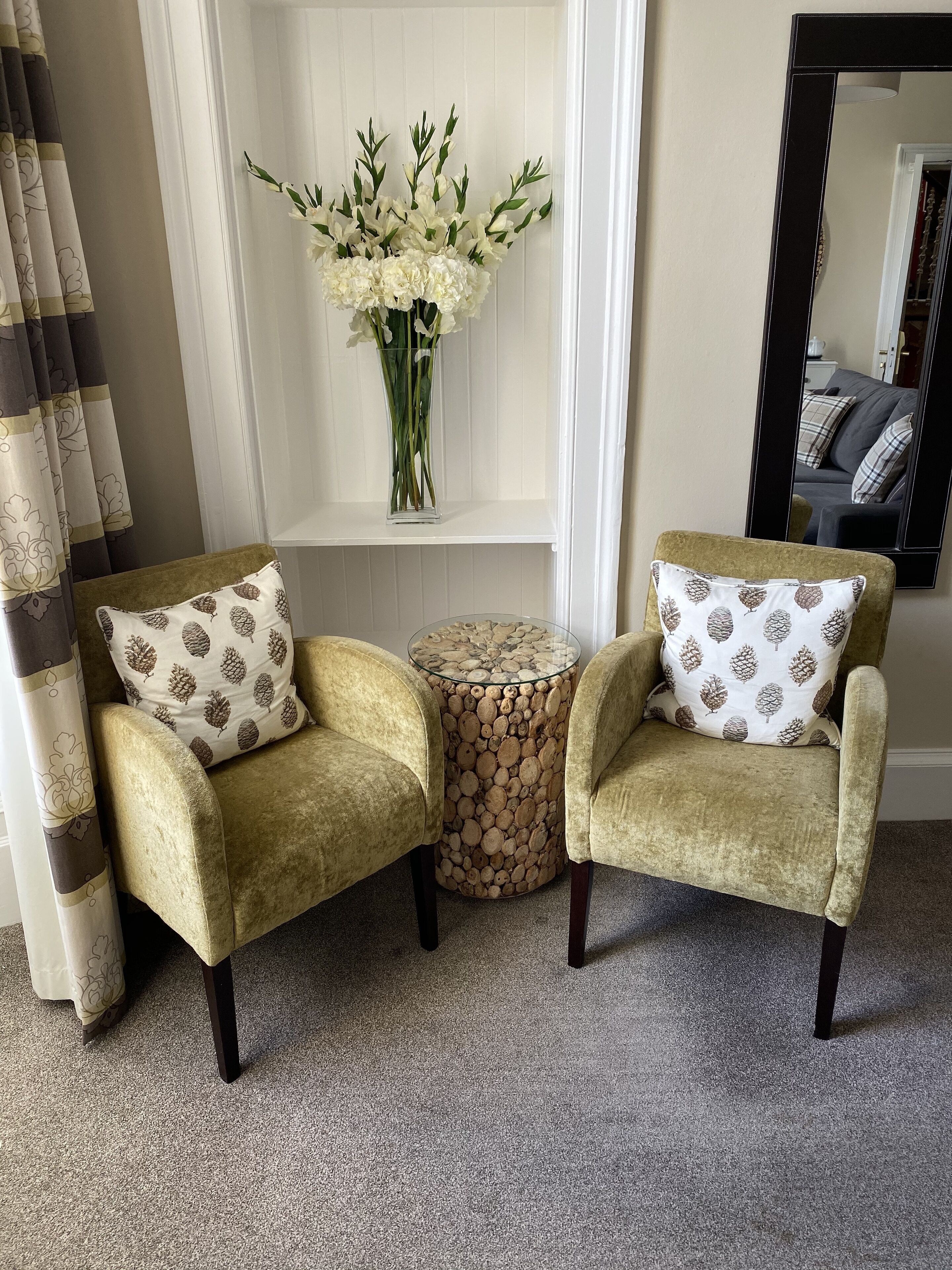 Two armchairs with patterned pillows, a small round table with a wood-like surface, and a vase of white flowers on a white shelf.