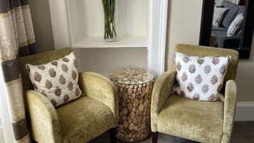 Two armchairs with patterned pillows, a small round table with a wood-like surface, and a vase of white flowers on a white shelf.