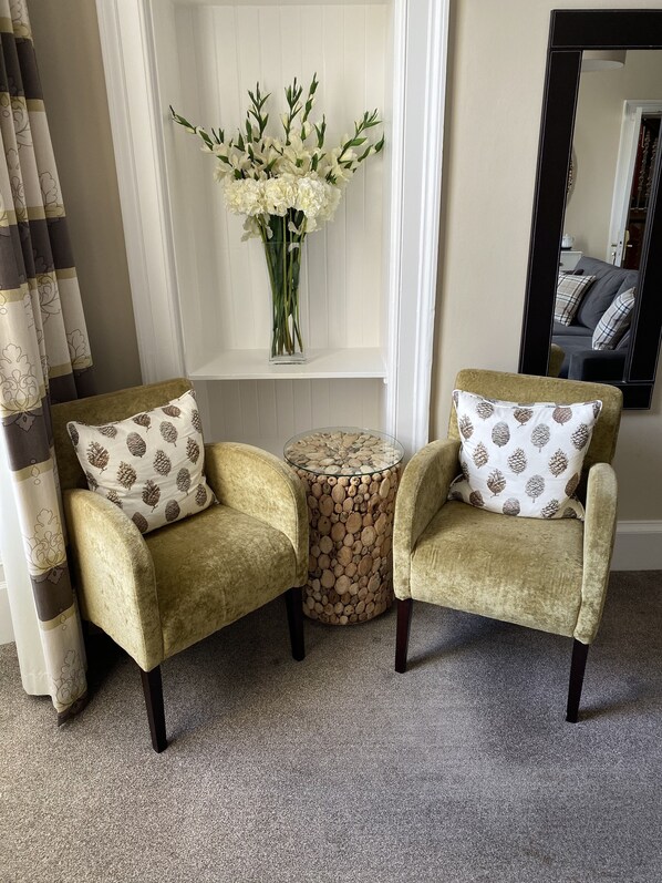 Dos sillones con cojines estampados, una mesita redonda con apariencia de madera y un florero con flores blancas sobre un estante blanco.