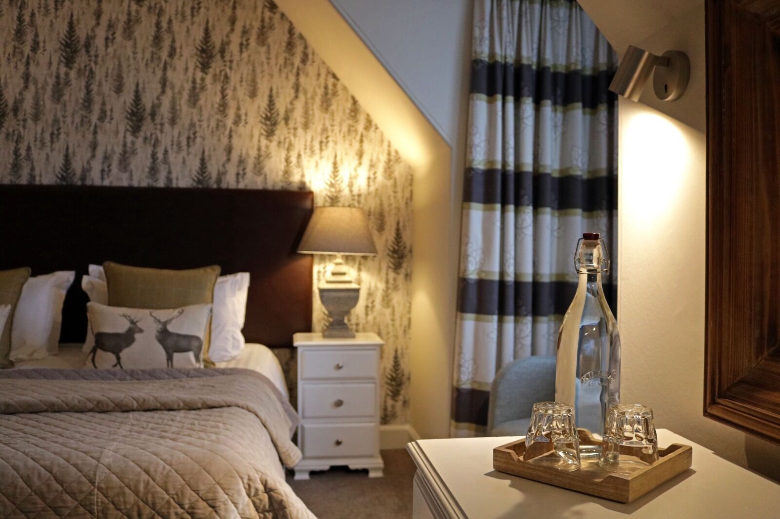Une chambre d’hôtel avec un lit, une table de chevet, une lampe, une bouteille et des verres sur un plateau.