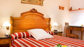 Habitación de hotel con cabecera de madera, colcha a rayas, dos lámparas de noche, un escritorio con silla y un estante con objetos decorativos.