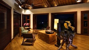 A spacious room with a high ceiling, wooden beams, a ceiling fan, a wooden table with chairs, and a large window with a view of the city at night.