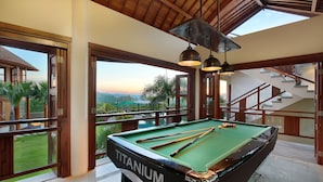 A pool table labeled "TITANIUM" in a spacious room with large windows, wooden beams, and a staircase.
