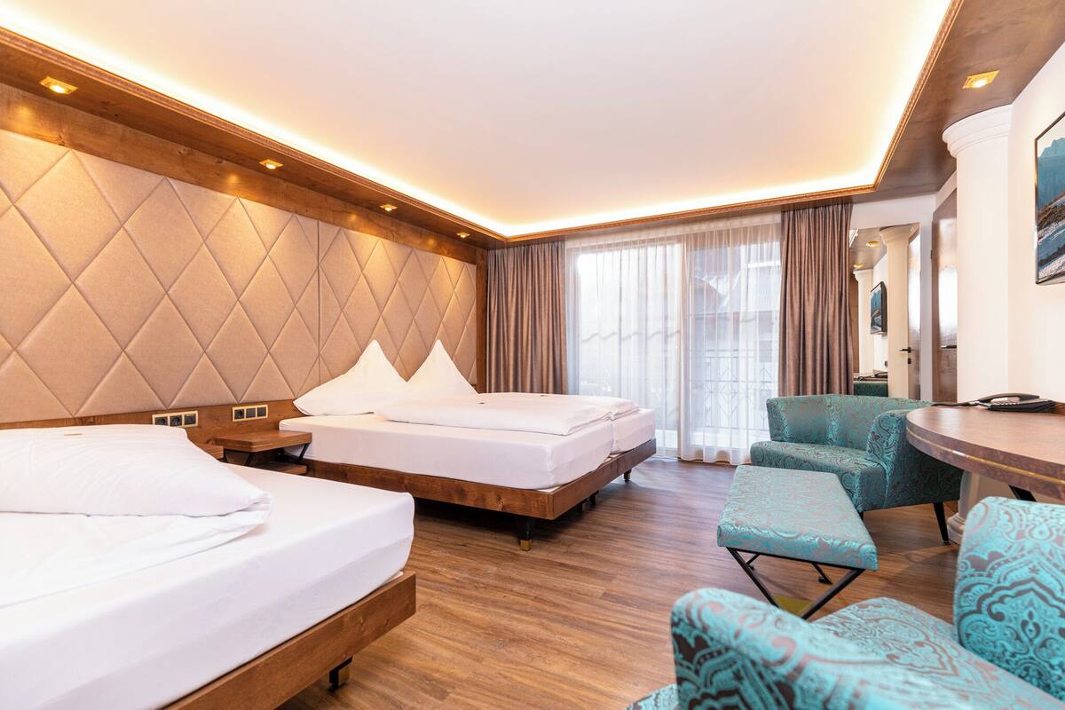 Habitación de hotel con dos camas, una zona de estar con una mesa y un amplio ventanal con cortinas.