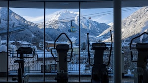 Ein Fitnessraum mit Trainingsgeräten und Blick auf schneebedeckte Berge sowie eine Seilbahn.