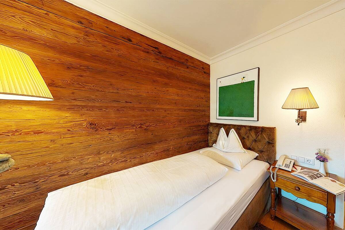 Habitación de hotel con cabecera de madera, cama con ropa de cama blanca, mesita de noche con lámpara y un cuadro en la pared.