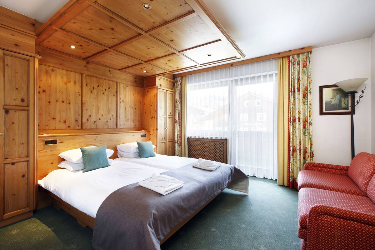 Habitación de hotel con techo de madera, una cama grande con ropa de cama blanca, un sofá rojo y una ventana con cortinas.